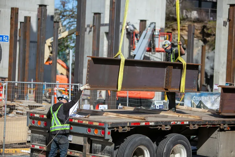 Crane lifting a steel beam off a delivery truck, guided by a worker on site
